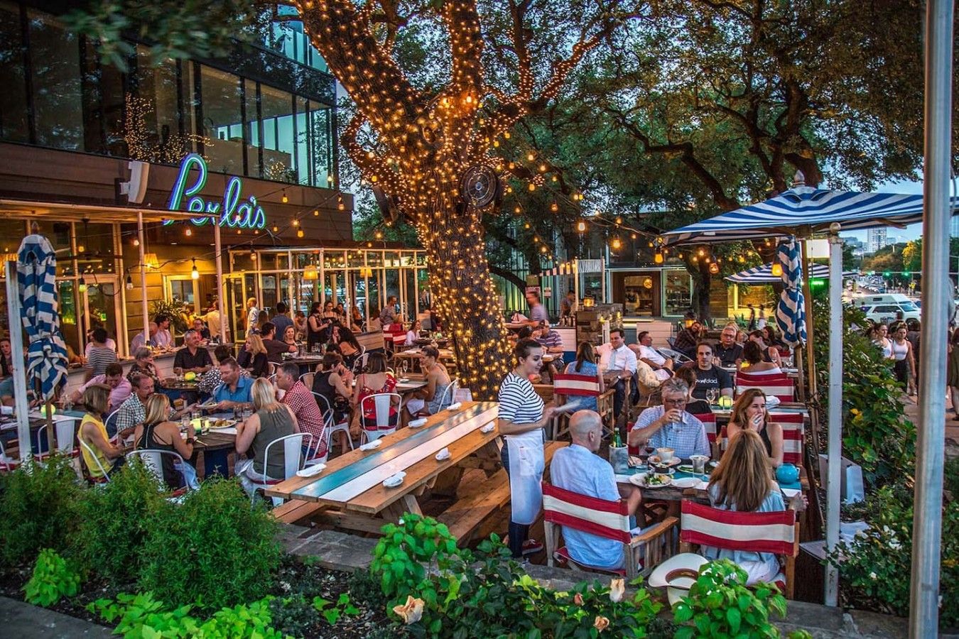 An outdoor dining area with tables and chairs, surrounded by greenery and a building with a neon sign.