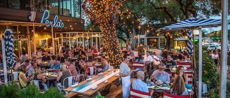 An outdoor dining area with tables and chairs, surrounded by greenery and a building with a neon sign.
