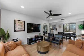 A modern living room with a flat-screen TV, comfortable seating, a dining area, and a large window offering a view.
