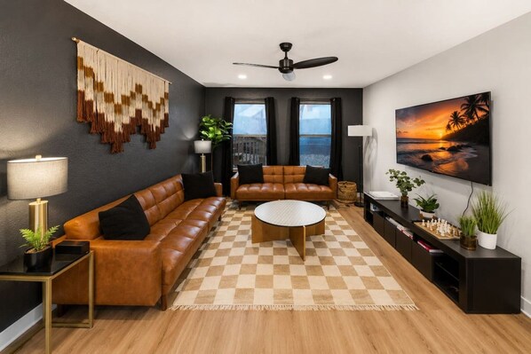 A modern living room with a checkered floor, leather sofas, a ceiling fan, a TV, and a painting of a sunset. - Austin Hideaway | 3BR Home with Private Backyard (Austin)