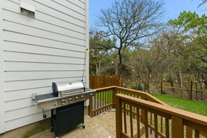 A barbecue grill on a porch with a wooden railing, a wooden fence, and trees in the background.