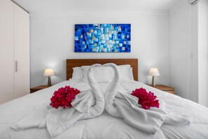 A hotel room with a neatly made bed, two bedside lamps, a wooden headboard, and a blue abstract painting on the wall. - Palm Aruba 418 | Modern Island Getaway (Palma Real)