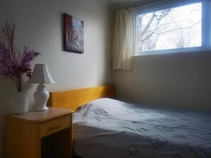 A room with a bed, a nightstand, a lamp, a vase with flowers, a painting on the wall, and a window with curtains. - A quiet and private small home where you can cook and do laundry. (Ottawa)