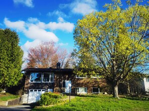 Una casa de dues plantes amb façana de maó, una porta de garatge visible i un gran arbre al jardí davanter.