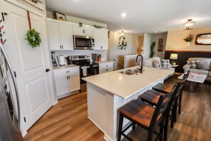 A modern kitchen with white cabinetry, a central island with bar stools, and a dining area with a sofa and coffee table. - 4 BR | 3 BA | New Build | 3-Car Garage | 5 Beds (Omaha)