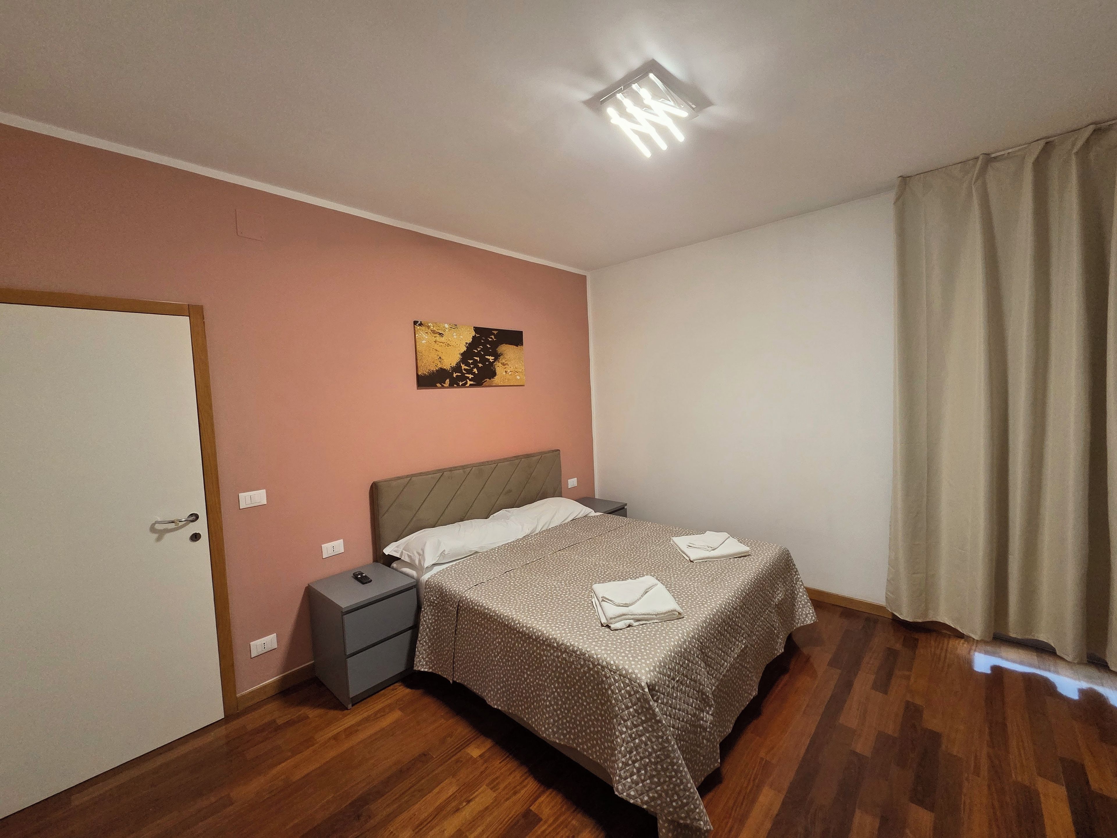Ein Schlafzimmer mit einem Bett, einem Nachttisch, einer Wandleuchte, einer Tür und einem Vorhang.