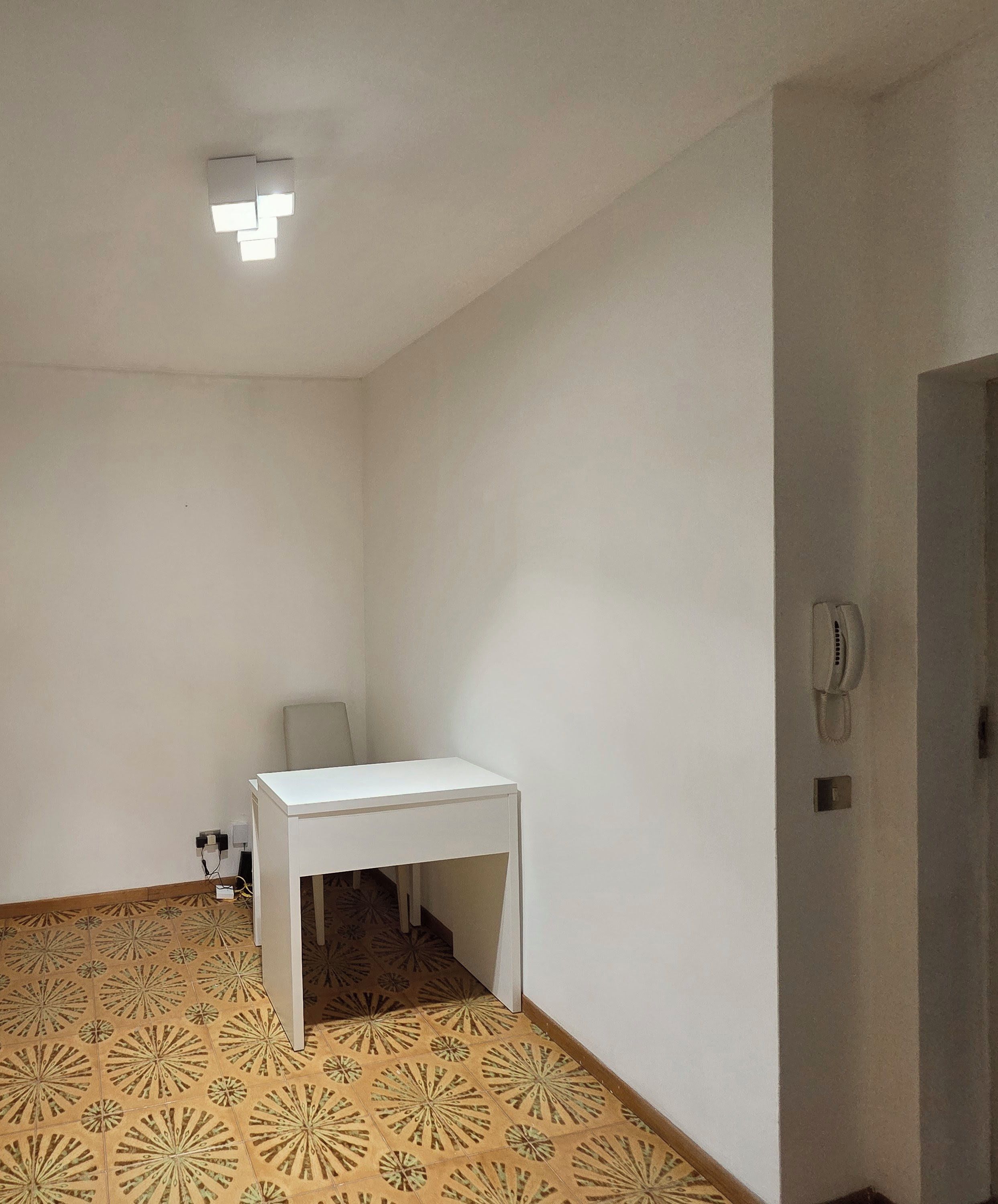 Una habitación con una mesa blanca, piso estampado, una silla, un teléfono y una lámpara.