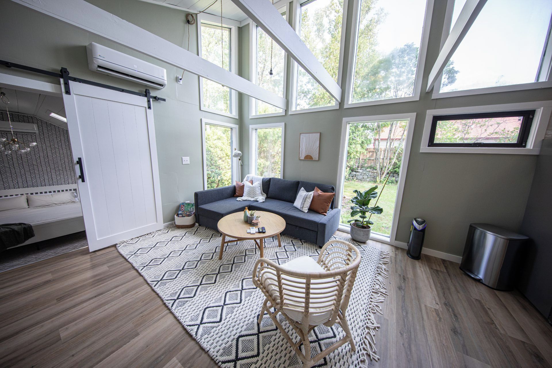 A room with a bed, a sofa, a round table, and a chair. There are windows, a plant, and a trash can.