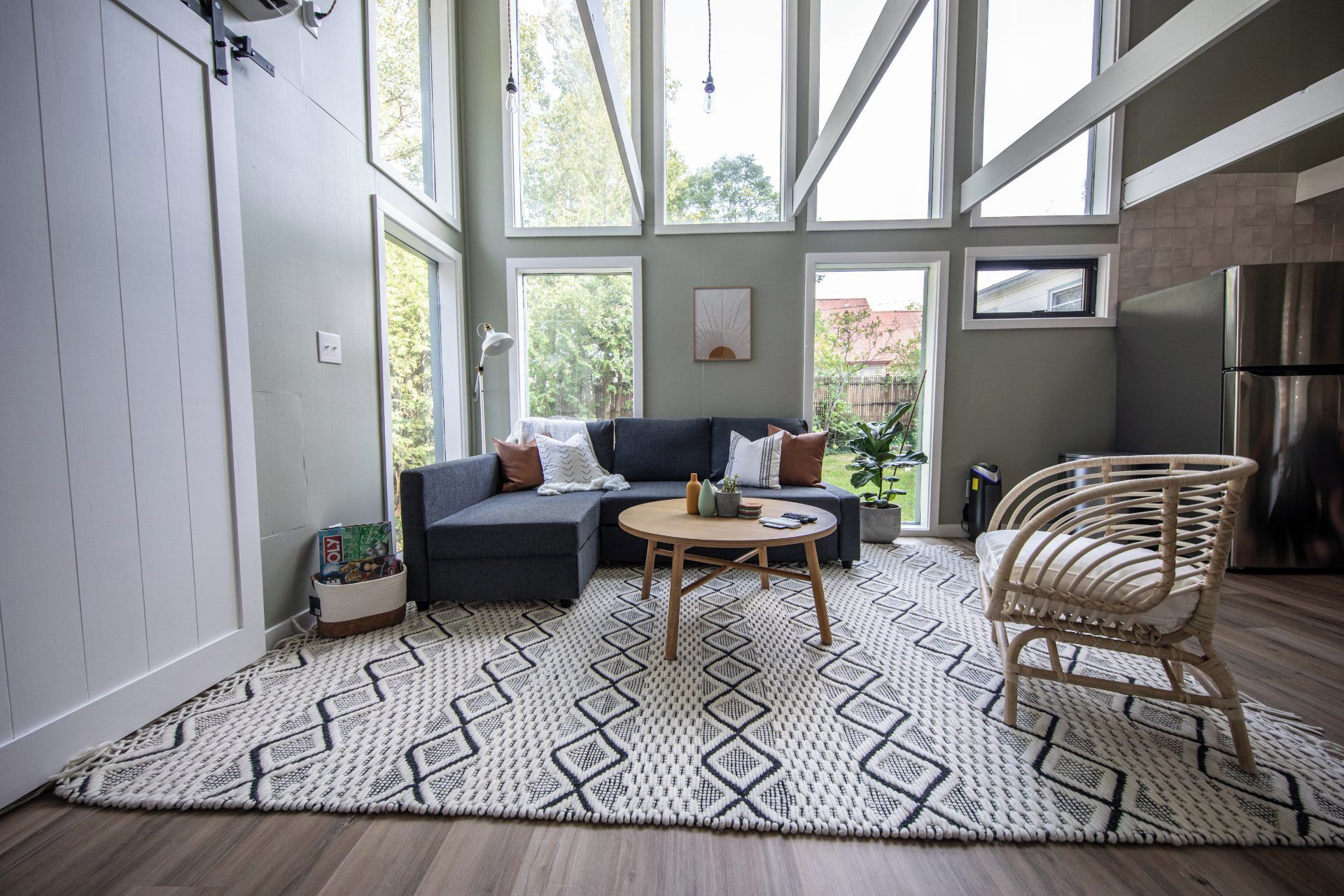 Un salon comprenant un canapé, une table basse ronde, un tapis à motifs, un fauteuil en osier et une grande fenêtre donnant sur un paysage verdoyant.