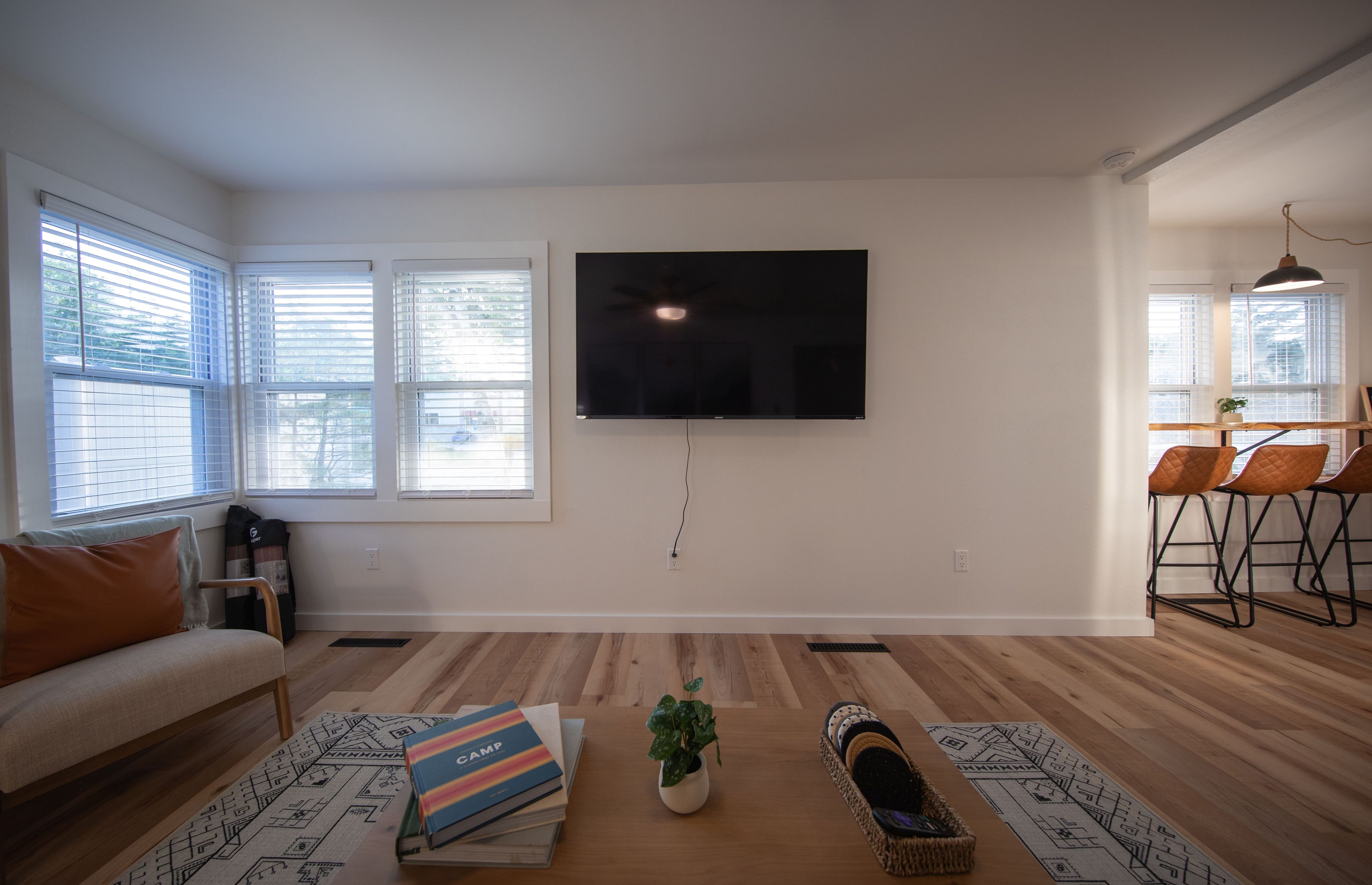 A living room with a flat-screen TV mounted on the wall, a sofa, a coffee table with books and a plant, and a dining area with chairs.