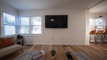 A living room with a flat-screen TV mounted on the wall, a sofa, a coffee table with books and a plant, and a dining area with chairs.