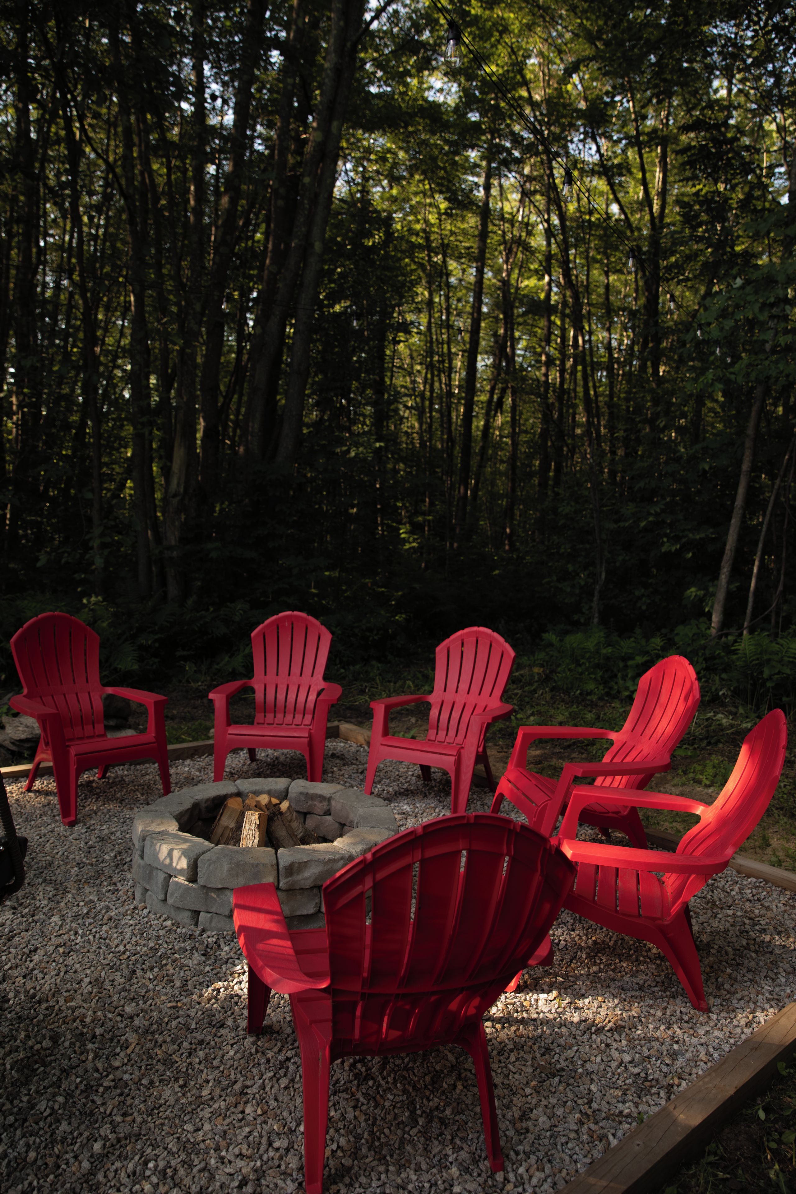 A fire pit with red chairs arranged around it, set on gravel with a forest backdrop.