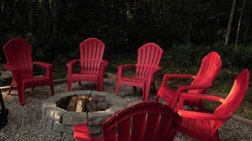 A fire pit with red chairs arranged around it, set on gravel with a forest backdrop.