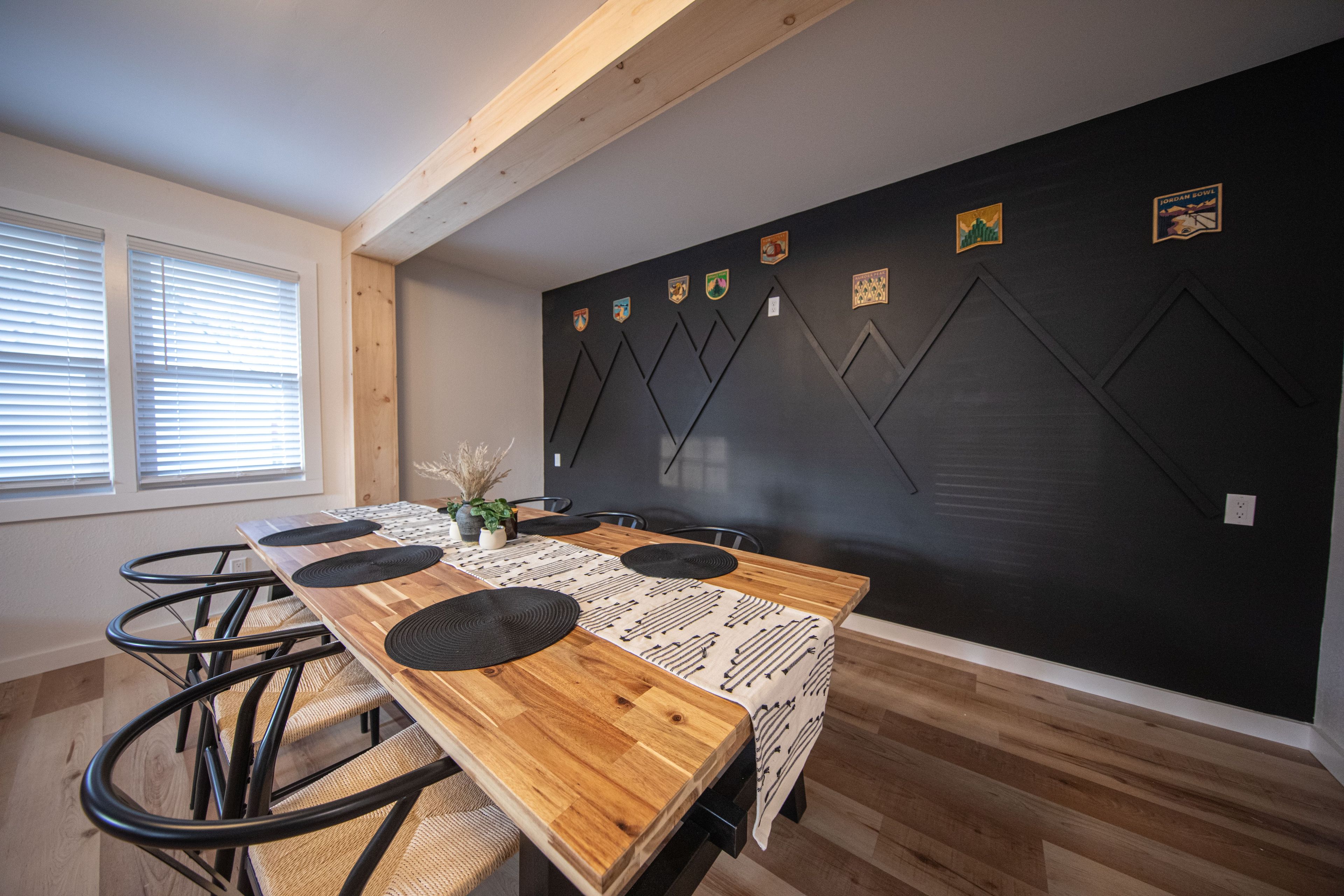 Un coin repas avec une longue table en bois, des sets de table noirs et des chaises. Il y a des fenêtres avec des stores sur le côté gauche.