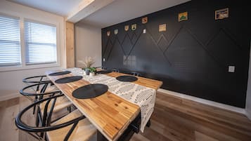 A dining area with a long wooden table, black placemats, and chairs. There are windows with blinds on the left.