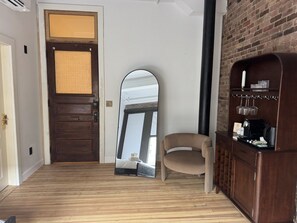 A room with a wooden floor, a brown door, a large mirror, a beige chair, and a wooden cabinet with a coffee machine. - Room 206, Ilium Luxury Hotel Downtown Troy (Troy)