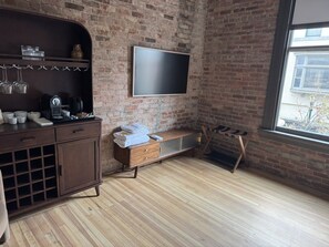 A room with a brick wall, a flat-screen TV mounted above a wooden bench, a wooden cabinet with wine glasses, and a window with a view of the outside. - Room 206, Ilium Luxury Hotel Downtown Troy (Troy)