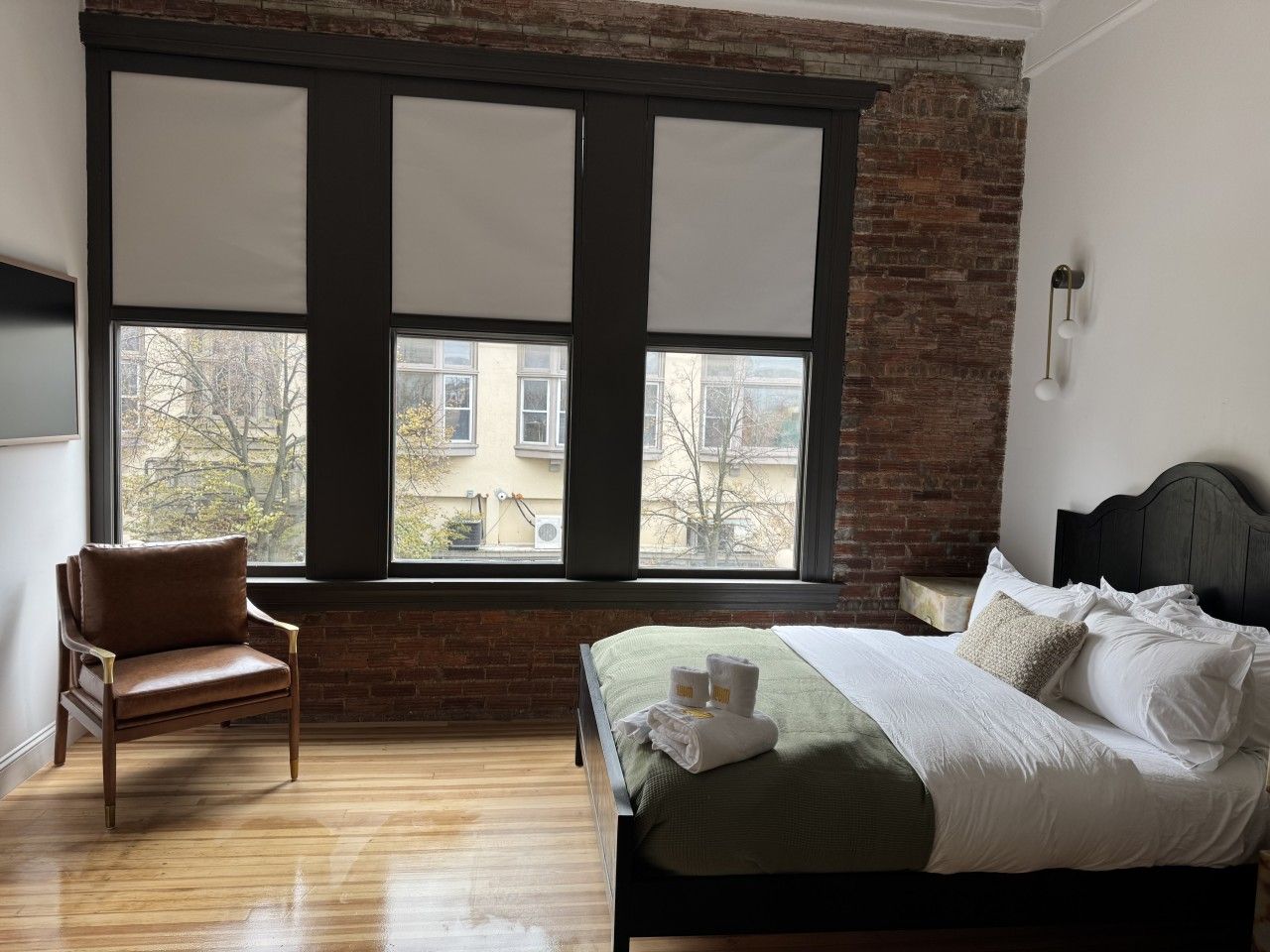 Habitación de hotel con un ventanal grande, cama con sábanas blancas, piso de madera y un sillón de cuero.