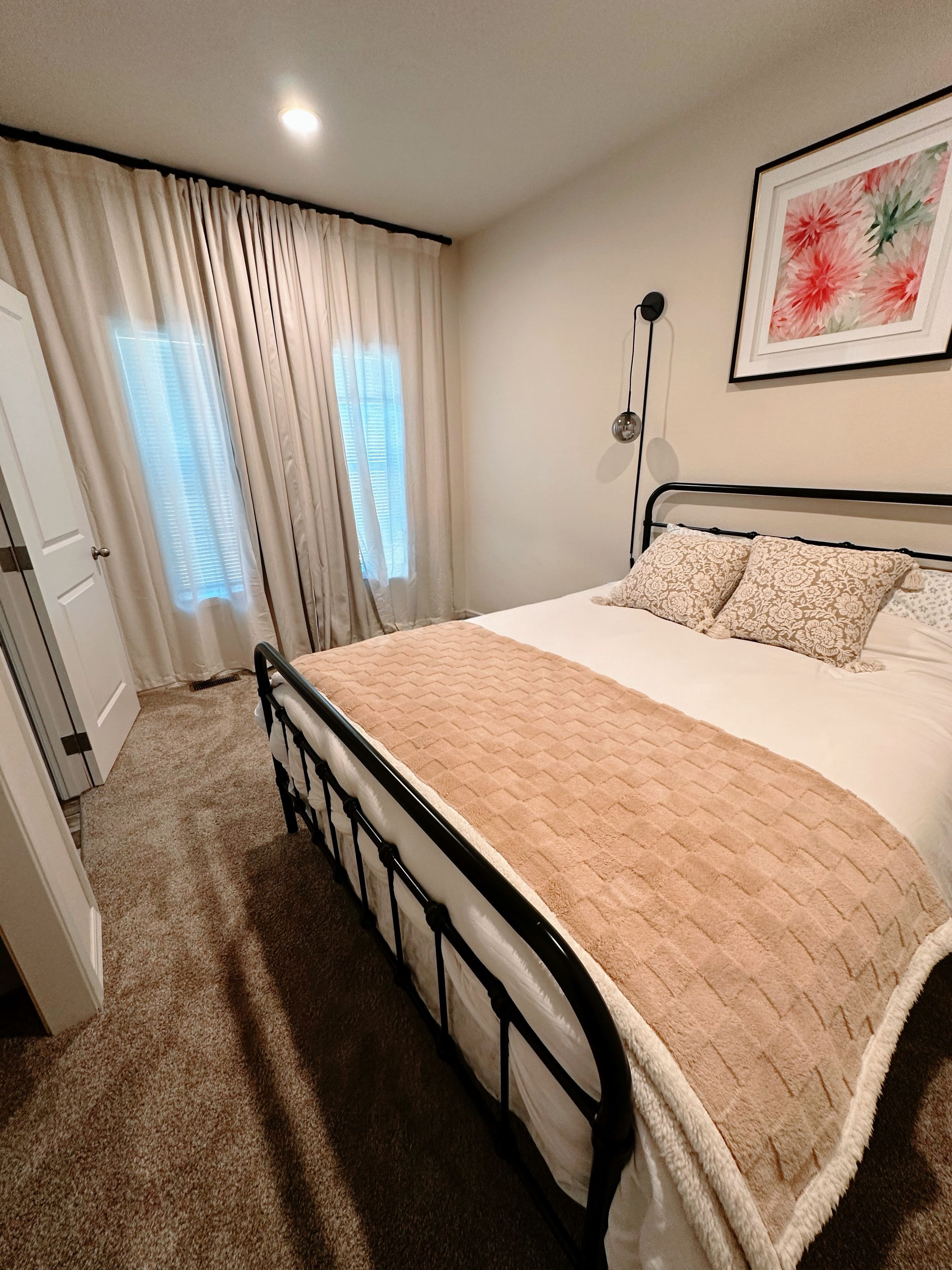 Une chambre à coucher avec un lit, des rideaux, une lampe fixée au mur et un tableau encadré.