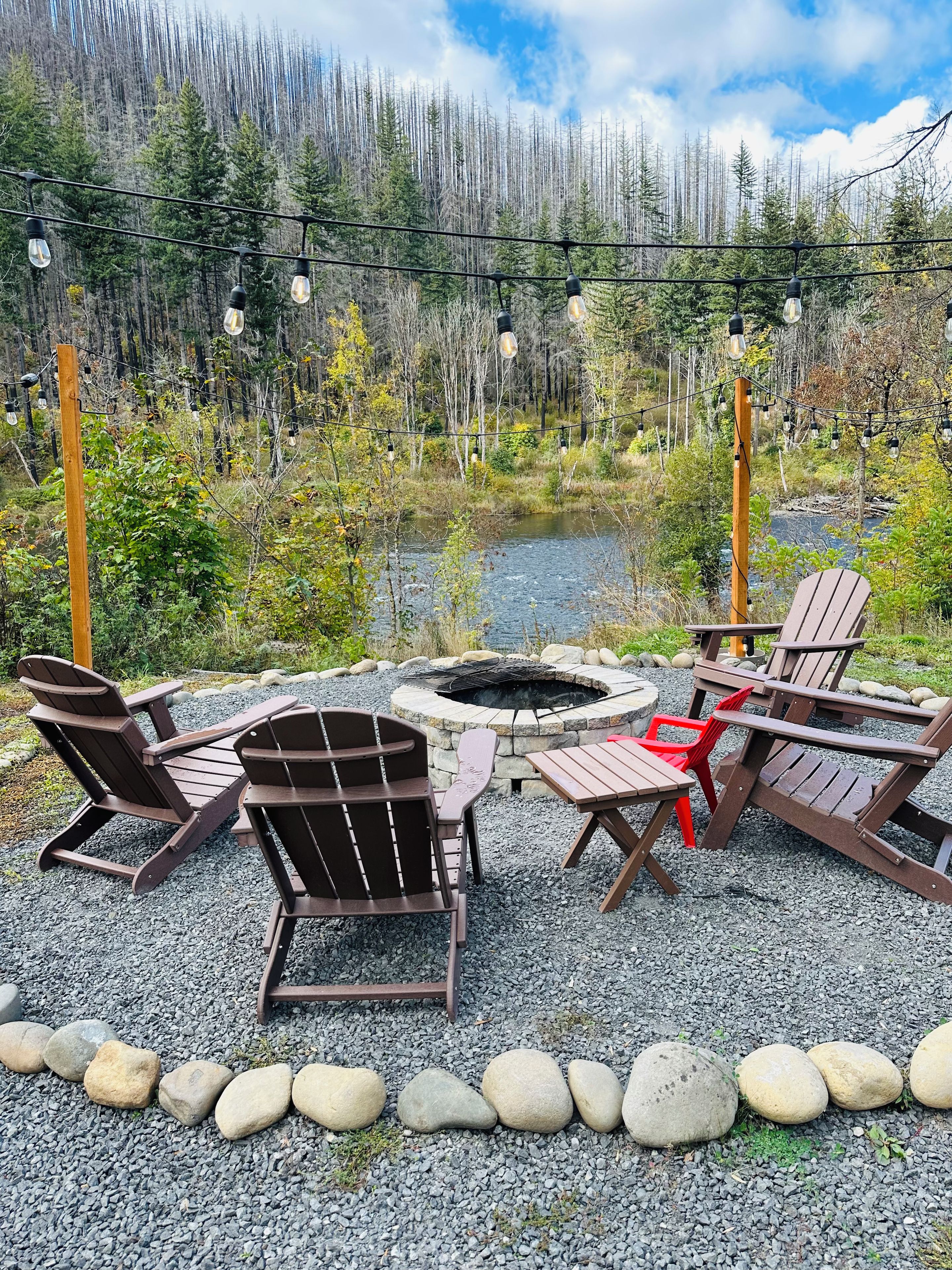 Un espace extérieur aménagé avec des chaises Adirondack et un foyer, entouré d’un chemin de gravier et bordé par une forêt.