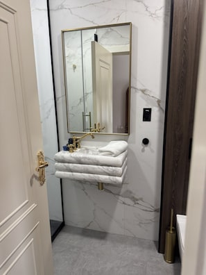 A bathroom with a marble wall, a gold-framed mirror, a white marble sink, and a gold toilet brush. - Room 202, Ilium Luxury Hotel Downtown Troy (Troy)