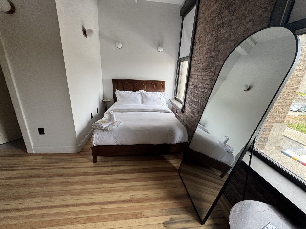 Un dormitorio moderno con suelo de madera, una cama con ropa de cama blanca, un espejo grande y vistas a la calle a través de una ventana.