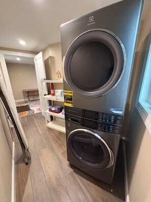 A compact, modern laundry room with a front-loading washer and dryer, a small shelf with cleaning supplies, and a patterned rug on the floor. - 3BR lakefront dog friendly home w/ views and kayaks (Colchester)