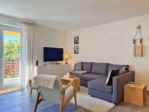Ein modernes Wohnzimmer mit einem grauen Sofa, einem Holzstuhl, einem Fernseher und einem kleinen Tisch mit einer Kerze.