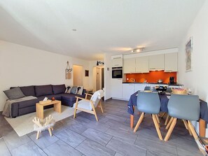 Ein modernes Wohnzimmer mit einer dunkelgrauen Couch, einem kleinen Holztisch und einem weißen Sessel. Im Hintergrund ist eine Küche mit weißen Schränken und einer orangefarbenen Spritzschutzwand zu sehen.