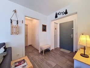 Ein modernes Wohnzimmer mit einer Couch, einem kleinen Tisch mit Büchern, einer Bank und einer Lampe. Es gibt zwei Türen, eine zu einem Flur und eine zum Schlafzimmer.