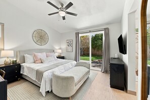 Een slaapkamer met een bed, nachtkastjes, een plafondventilator, een groot raam met gordijnen en een aan de muur bevestigde televisie.