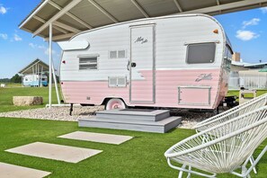 A pink and white RV parked under a covered area with steps leading up to it, surrounded by a grassy lawn and a white chair. - Blue Skies Resort | Glamping | Pool | Fireplace (Fredericksburg)