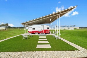A recreational vehicle (RV) under a covered area with a paved walkway leading to it, surrounded by grass and gravel. - Blue Skies Resort | Glamping | Pool | Fireplace (Fredericksburg)