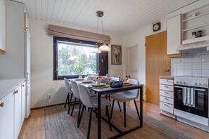 A dining area with a table set for four, a modern kitchen with a stove, and a window with a view of greenery. - Cozy Family House in the heart of everything (Rovaniemi)