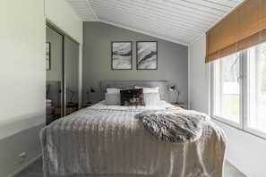 Ein Schlafzimmer mit einem Bett, Nachttischen, Lampen, einem Fenster mit Jalousien und zwei gerahmten Bildern an der Wand.
