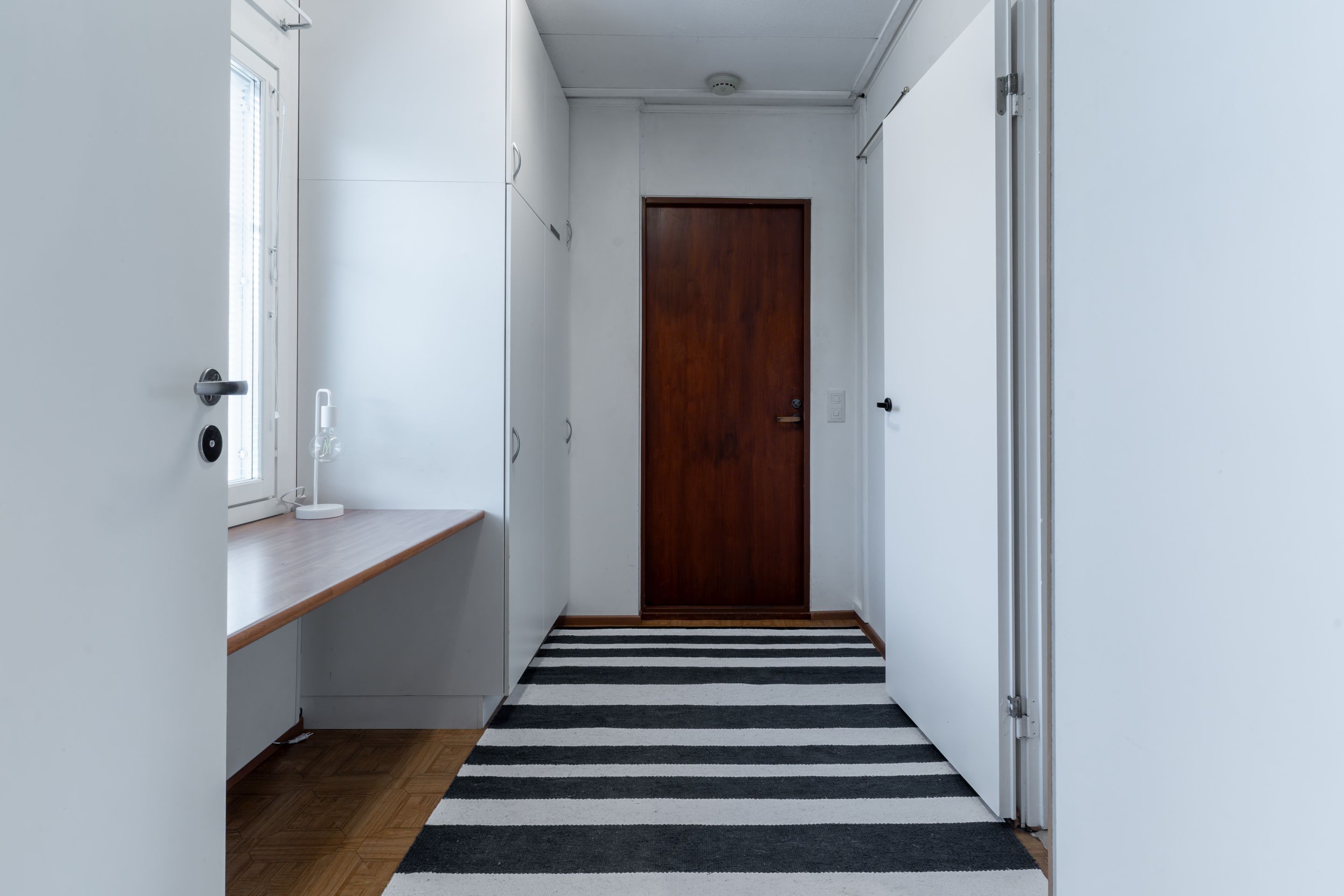 Un pasillo con puerta de madera, paredes blancas, un banco y un felpudo a rayas.
