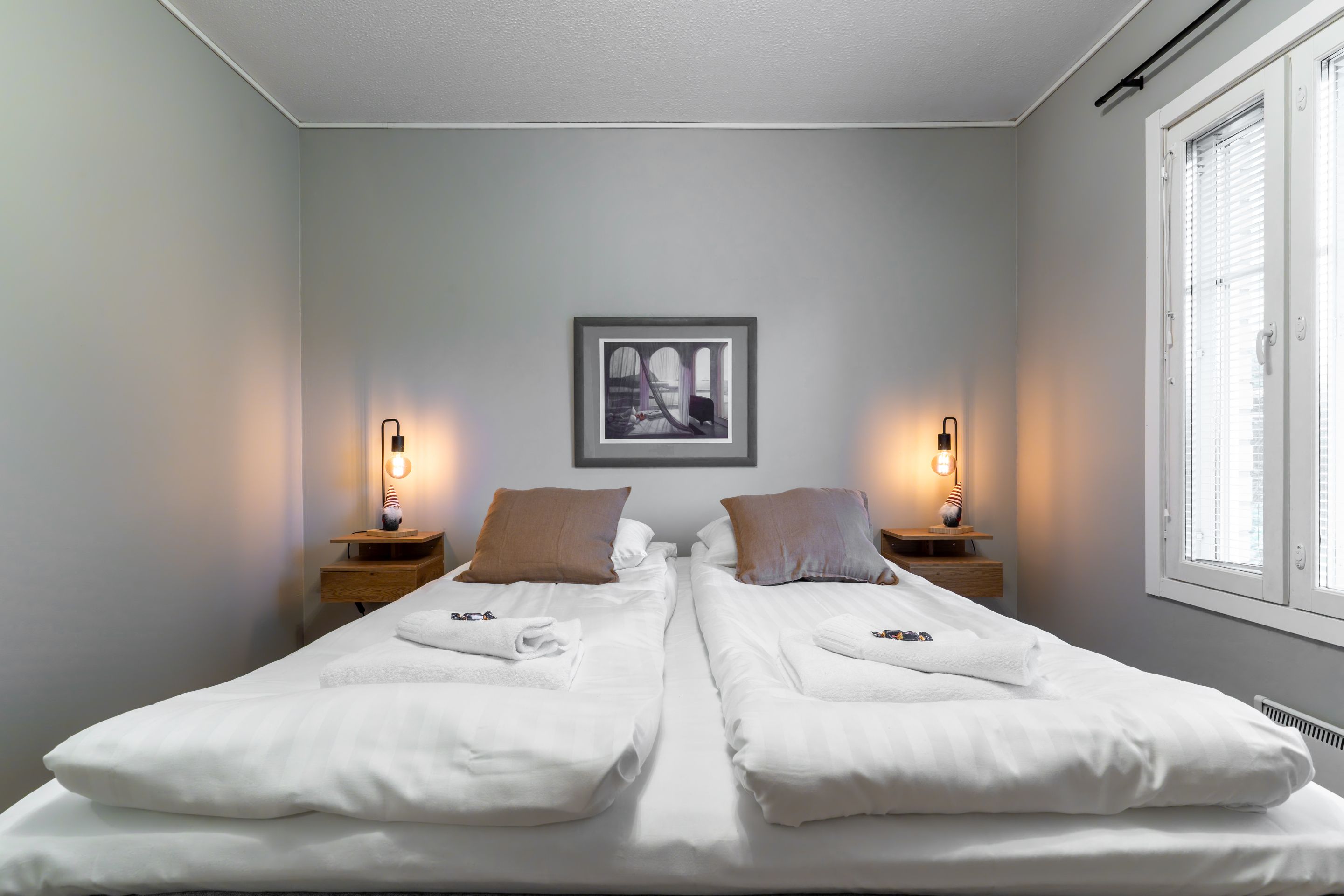 A double bed with white linens, two bedside tables with lamps, a framed picture on the wall, and a window with blinds.