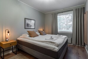 A modern bedroom with a large bed, a bedside table with a lamp, a framed picture on the wall, and a window with curtains. - Villa Santa's Star I near SCV (Rovaniemi)