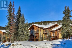 Un chalet à étages, dont le toit est recouvert de neige, entouré de grands arbres à feuilles persistantes.