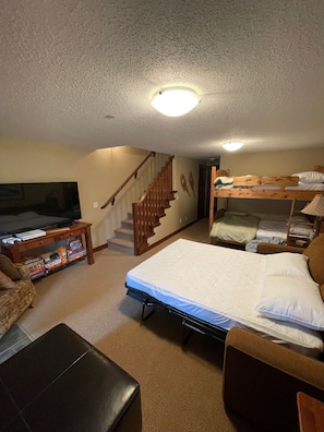 A compact room with a bunk bed, a flat-screen TV, a wooden table with board games, a staircase, and a couch. - Woodhaven #34 | 3 Peaks Rentals (Sun Peaks)