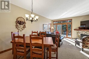 A dining area with a wooden table and chairs, a chandelier, a fireplace, and a large window with a view of trees. - Woodhaven #34 | 3 Peaks Rentals (Sun Peaks)