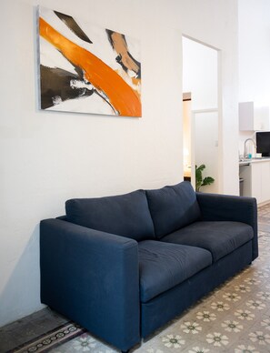 A modern living room with a dark blue sofa, a large abstract painting, and a kitchen area in the background. - Nando's Place 1 - Charming 1BR Apt near Old San Juan and Beach (San Juan)