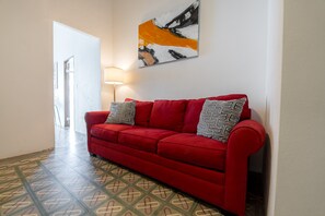 Un canapé rouge avec des coussins à motifs, placé dans un couloir dont le sol est également à motifs, avec une lampe et un tableau accroché au mur.