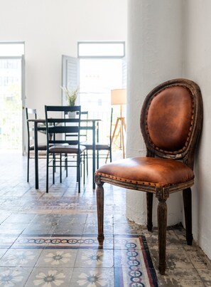 A chair with a cushioned seat and wooden legs, positioned against a wall in a room with patterned flooring and a table with chairs in the background. - Nando's Place 4 - Charming 1BR Apt near Old San Juan and Beach (San Juan)