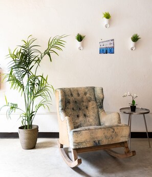 A rocking chair with a tufted backrest, a potted plant with long leaves, a small round table with a potted orchid, and a wall-mounted picture frame. - Juliette Hostel, Bed in 8-Bed Female Dorm, Old San Juan (San Juan)