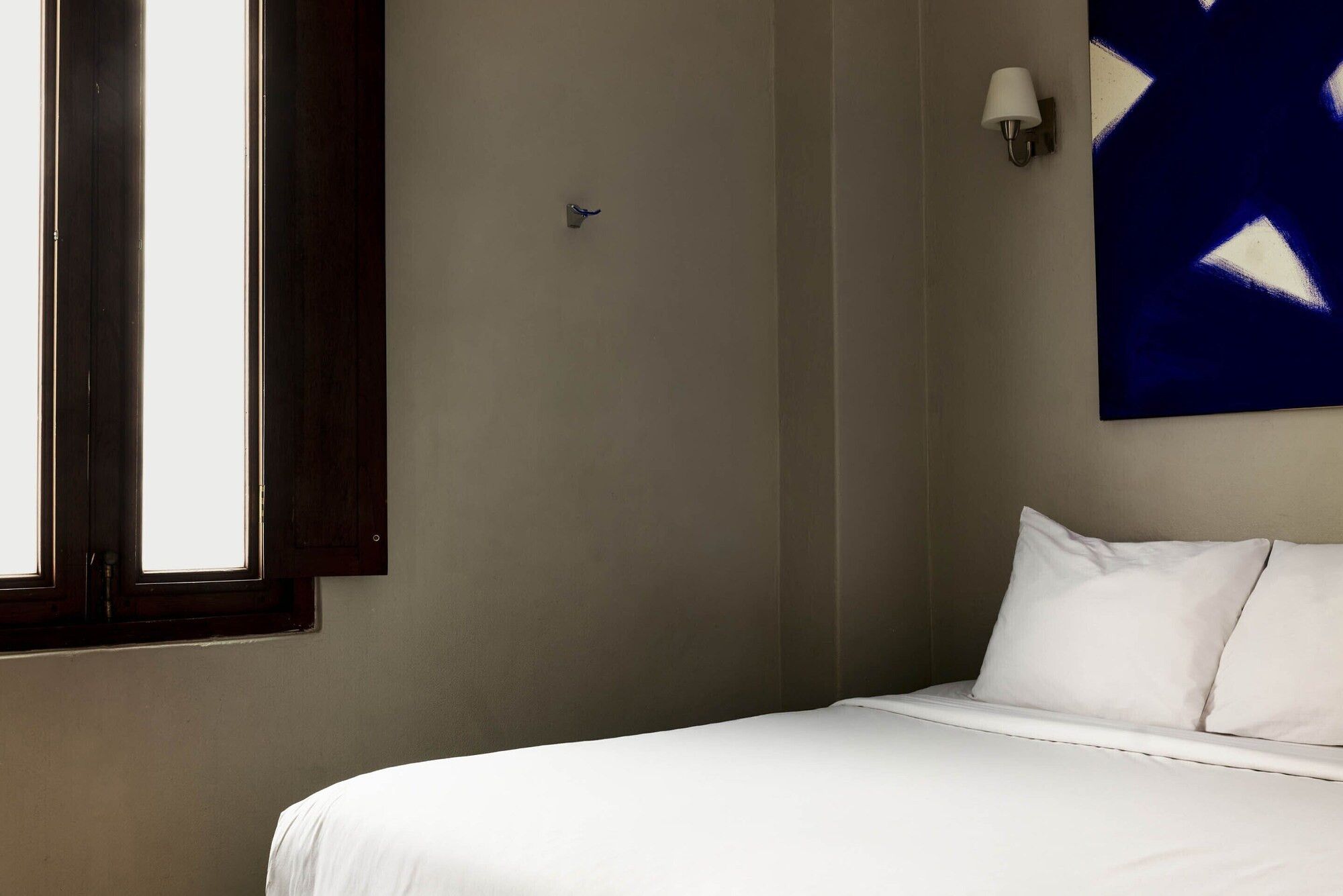 A hotel room with a bed, a window, a wall-mounted light, and a blue abstract painting.
