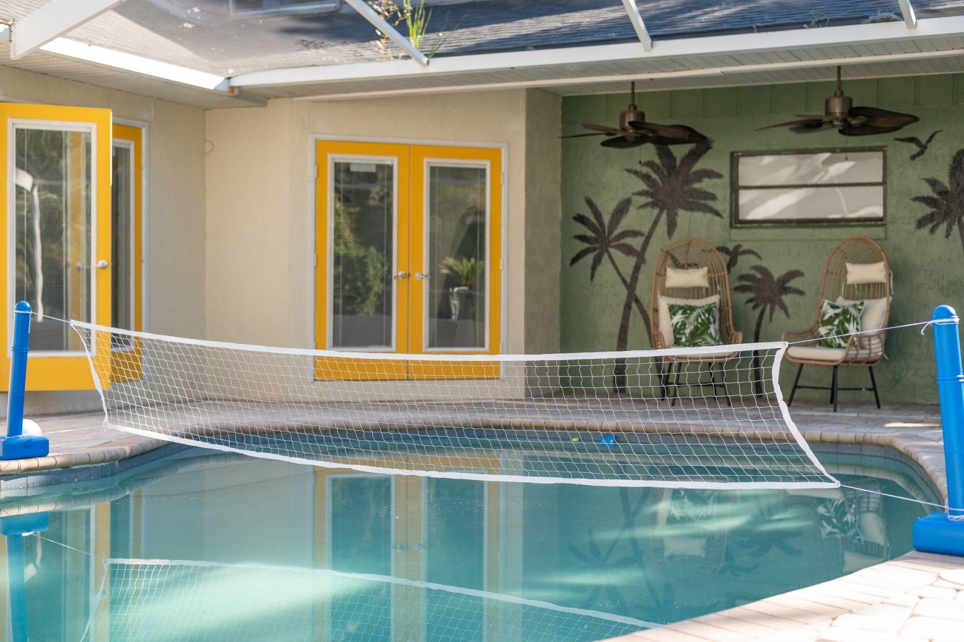 Una piscina con zona delimitada por una red, una puerta con paneles de vidrio y un muro con murales de palmeras.