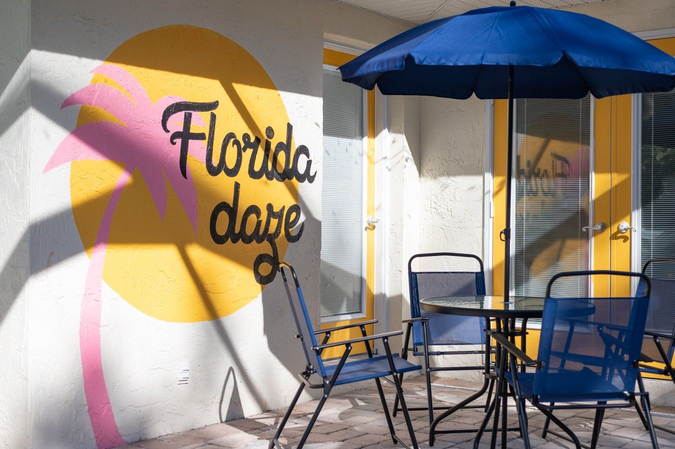 Un patio con mesa y sillas, un paraguas azul, y una pared con un árbol de palma pintado y la inscripción 'Florida dear'.