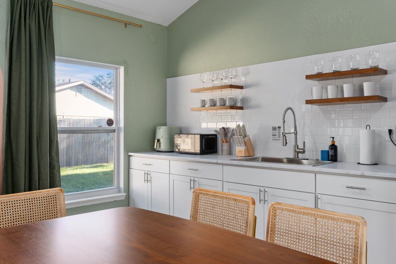 Una cocina con armarios blancos, una mesa de comedor de madera, sillas de mimbre y vistas a un patio trasero a través de una ventana.