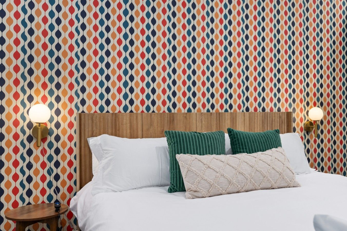 Una cama con ropa de cama blanca, dos almohadas verdes y un cabecero de madera. Hay dos lámparas de pared a ambos lados de la cama.